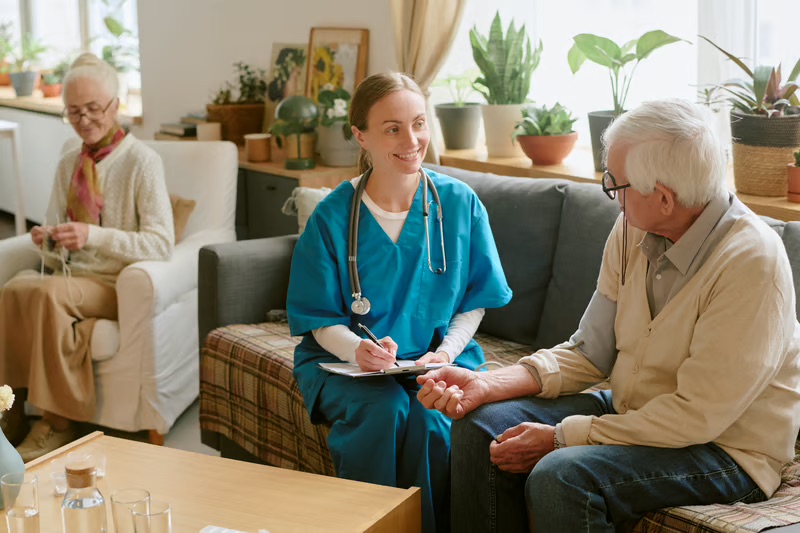 Hospice nurse speaking with an older patient during a home care visit while a family member sits nearby.